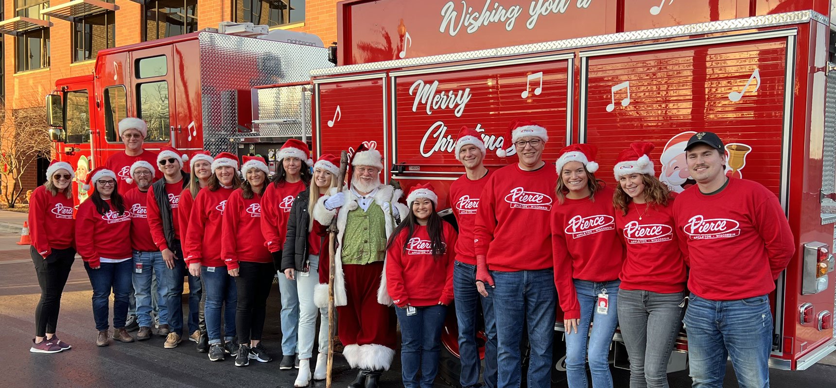 A group of Pierce employees stand with Santa in front of a firetruck.