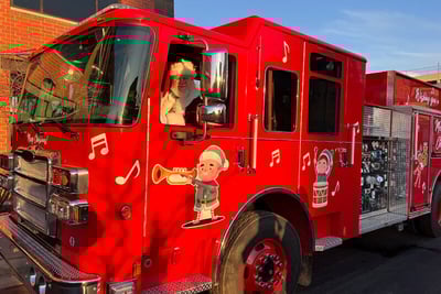 Santa sits in the driver seat of a holiday themed red Pierce fire truck. 
