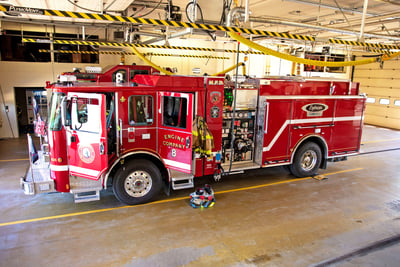 A red Pierce electric fire truck is parked inside a fire station plugged into an overhead charging station. 