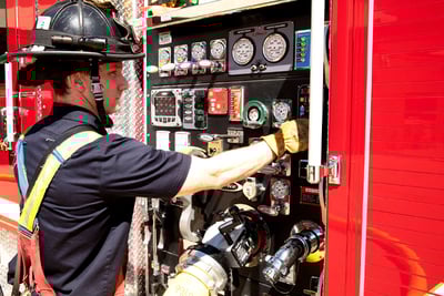 A firefighter in turnout pants, a navy-blue station shirt and a helmet works on an exterior fire truck panel.