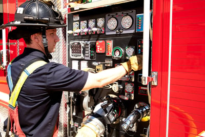 A firefighter in turnout pants, a navy-blue station shirt and a helmet works on an exterior fire truck panel.