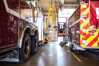 A red electric fire truck is plugged into power in a fire station bay next to another red fire truck.