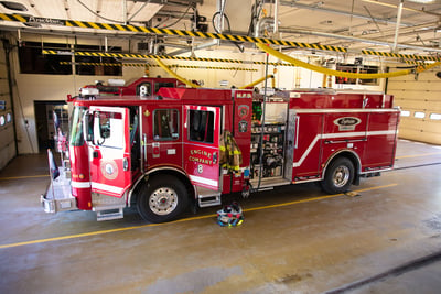 A red electric fire truck is parked in a fire station bay connected to an overhead power supply.