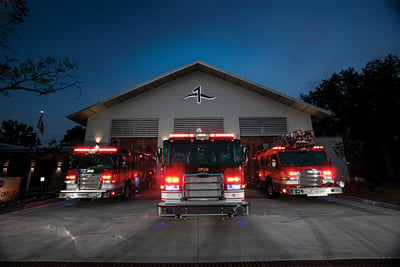 A fleet of vehicles built using a fire apparatus configurator tool are parked at dusk outside of a fire station with emergency lights turned on. 