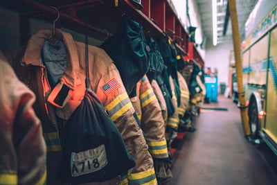 The interior of a fire station shows a row of lockers and hanging gear across from a parked Pierce fire truck.