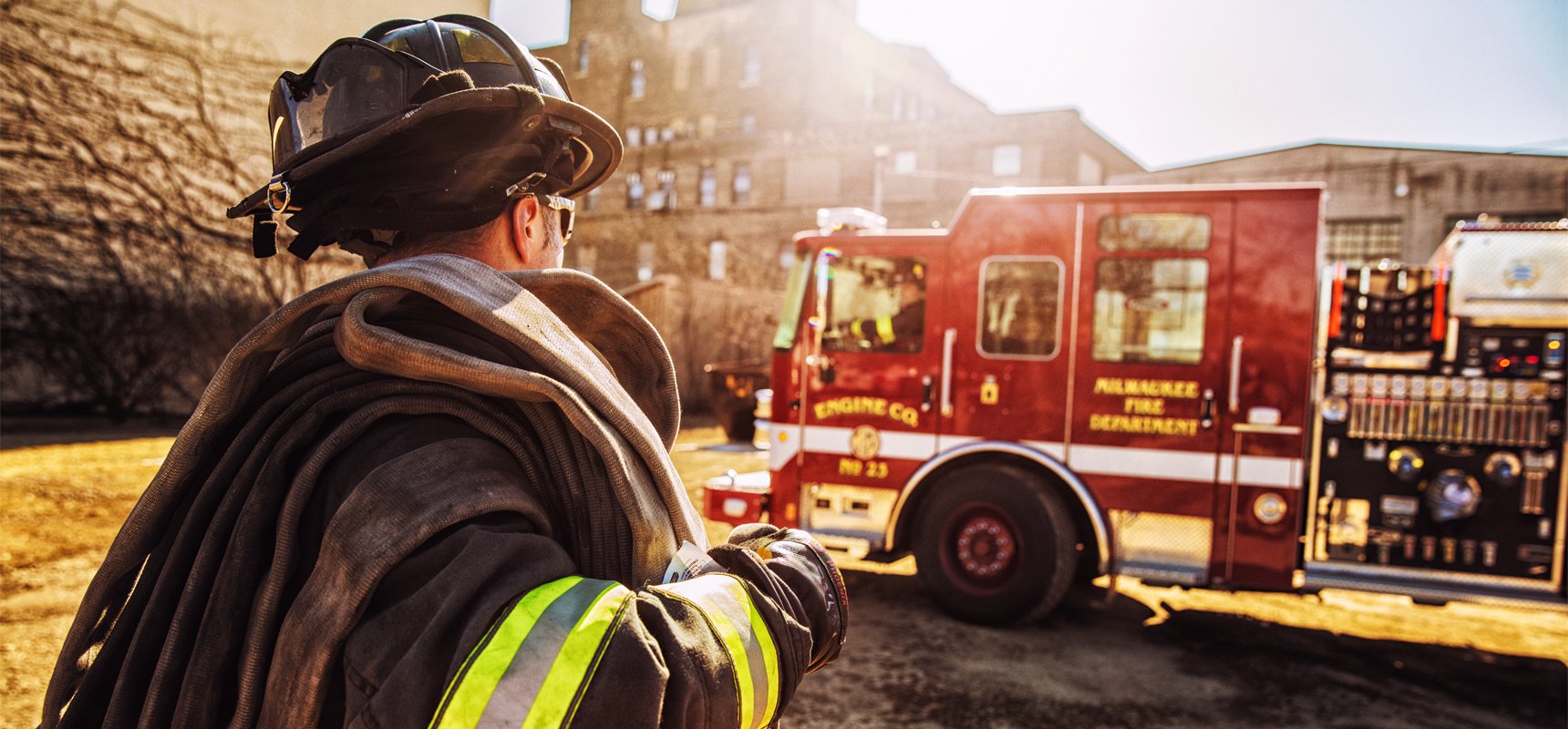A firefighter stands with a hose on his shoulder as he talks with the operator of a red fire truck in the background.