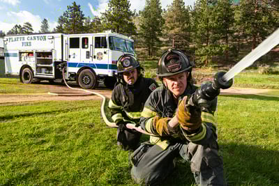 Two firefighters are holding a nozzle and hose as water sprays from a Pierce pumper fire truck. 