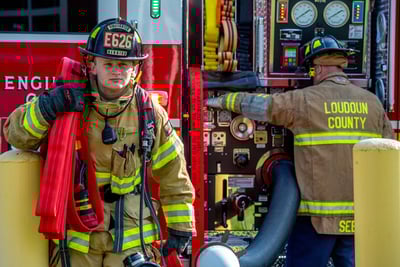 A firefighter is walking with hose on his shoulder as another operates the panel on an adjacent Pierce fire truck.