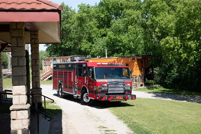 A red and black fire truck approaches a building structure driving on grass and gravel with portable construction buildings in the background. 