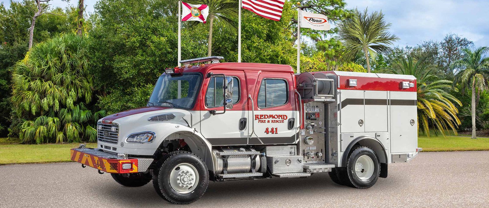 The white and red Redmond Fire & Rescue wildland pumper fire truck is parked on asphalt with flags and trees in the background. 