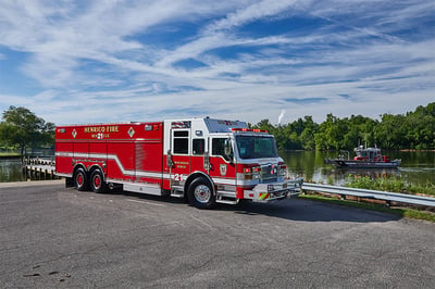 A red and white fire truck is parked on asphalt at the side of a river in a rural setting with a small watercraft in the background. 