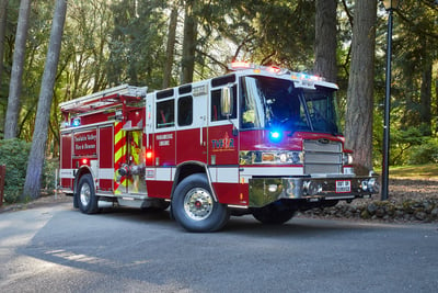A red and white fire truck from a rural fire department fleet drives uphill on an asphalt road in a wooded area.