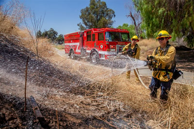 Two firefighters spray water at a brush fire on the side of a rural roadway with a red fire truck parked in the background.