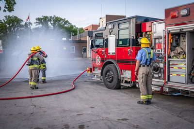 Two rural firefighters are spraying water at the back of a building while another firefighter manages the operators panel on the driver’s side of the red and black fire truck. 