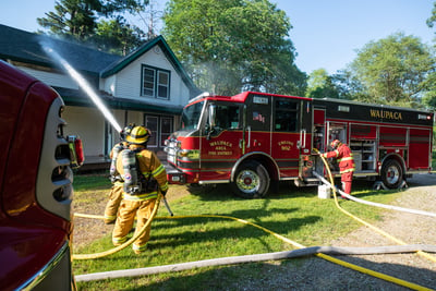 A red and black rural fire truck is parked in grass next to a farmhouse as firefighters in turnout gear spray water at the home. 