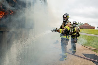 In a smoky environment, two firefighters hold a fire hose with a straight tip nozzle and aim water at a fire in the foreground.