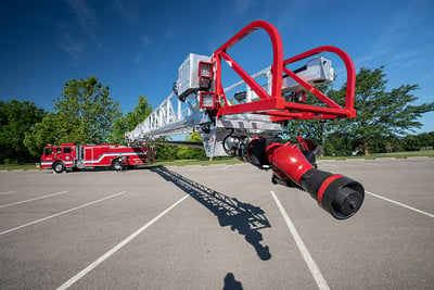 A red fog nozzle is affixed to an aerial ladder device extending out from a parked red fire truck in a parking lot. 