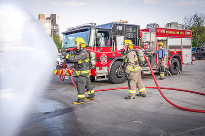 Two firefighters in full gear next to a red fire truck hold a fire hose with a fog nozzle as is sprays a wide-casting net of water in a parking lot. 