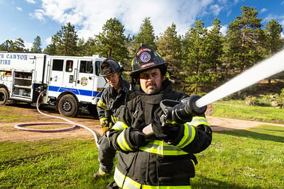 Two firefighters in a wooded area hold a fire hose with a straight tip nozzle as it shoots water from a white fire truck with a blue stripe.