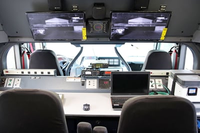 The interior of a hazmat fire trucks shows a command center featuring two desk stations with large overhead monitors, a hardwired computer and chairs. 