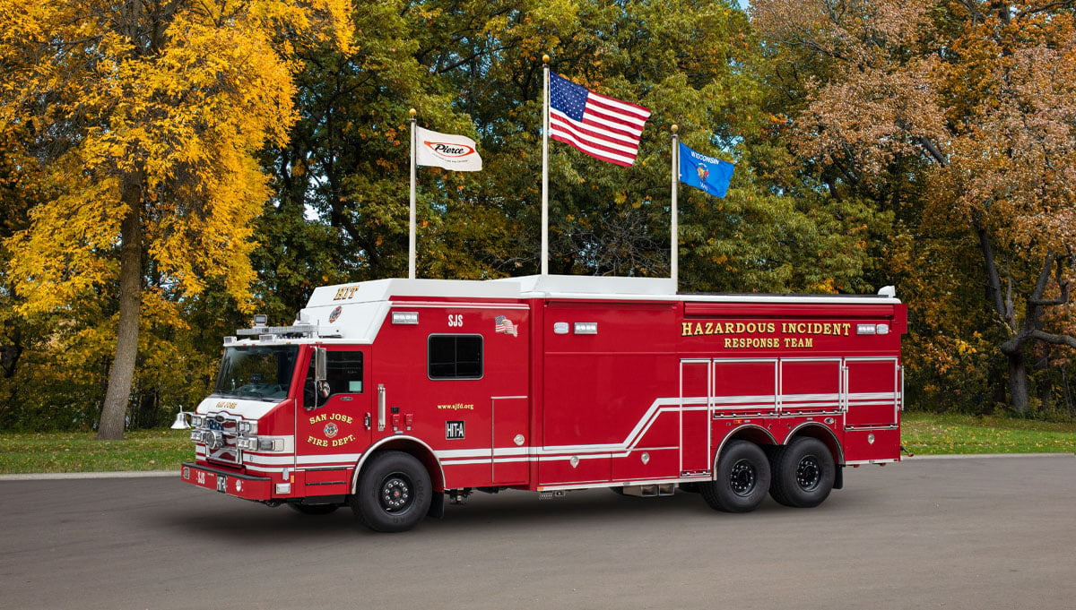 The red San Jose Fire Department combination hazmat rescue apparatus is parked outside in front of three waving flags and an autumn background. 