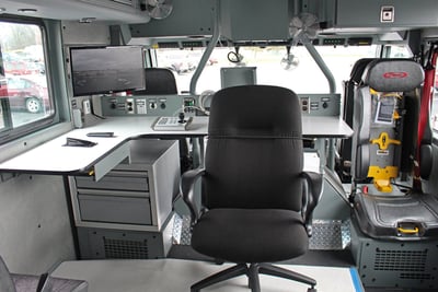 The interior of a hazmat fire truck shows a command center area, featuring a chair on rollers, a large desk and a several monitoring systems. 