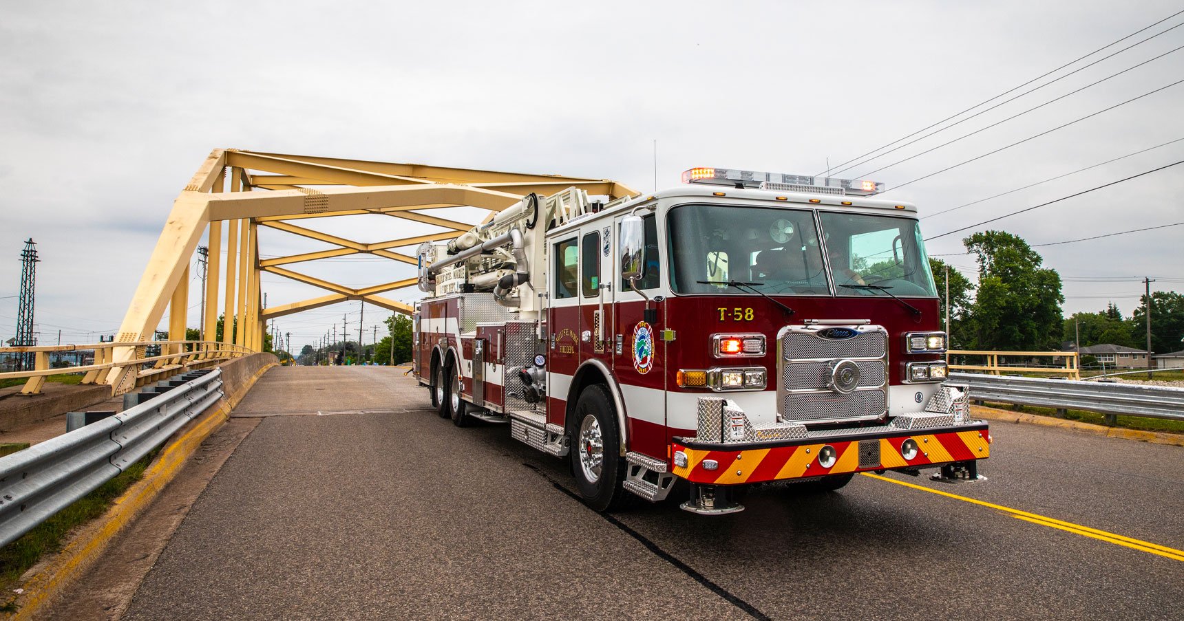 A red and white aerial fire truck designed for urban fires drives under a yellow steel bridge on an asphalt road.