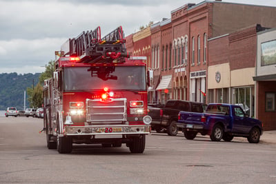 A red aerial ladder fire truck is driving down an asphalt road with parked cars and two-story shops lining one side of the street. 