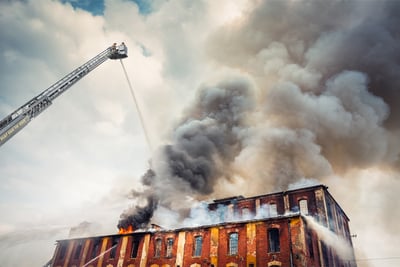 A dramatic photo shows one of the challenges of urban firefighting; an old brick building is aflame as an elevated aerial platform fire truck sprays water down, sending plumes of smoke into the sky. 