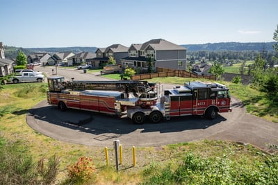 A red and black tiller apparatus is turning in a cul du sac in a residential block with greenery in the foreground and houses in the background. 