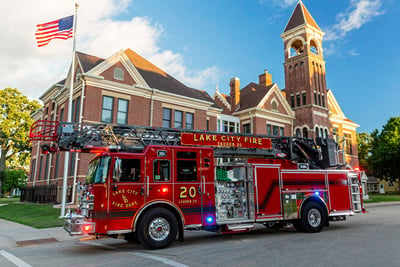A red urban aerial fire truck is driving on an asphalt road in front of a building with a bell tower and a blowing American flag. 
