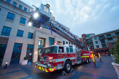 A red and white urban aerial platform fire truck extends its ladder with scene lights turned on in a condo complex at dusk.