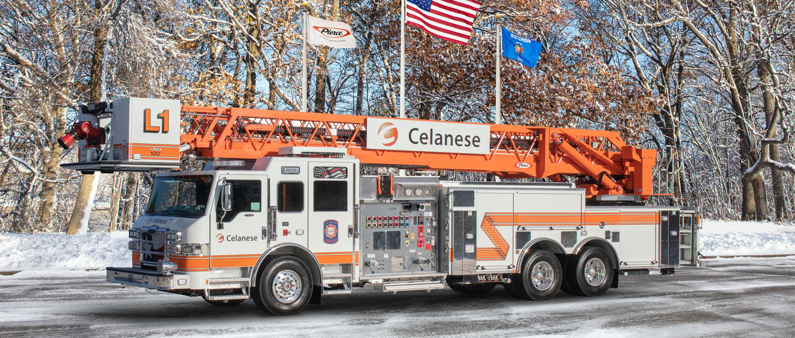 A white and orange aerial fire truck is parked in front of flags in the Pierce Manufacturing lot. 