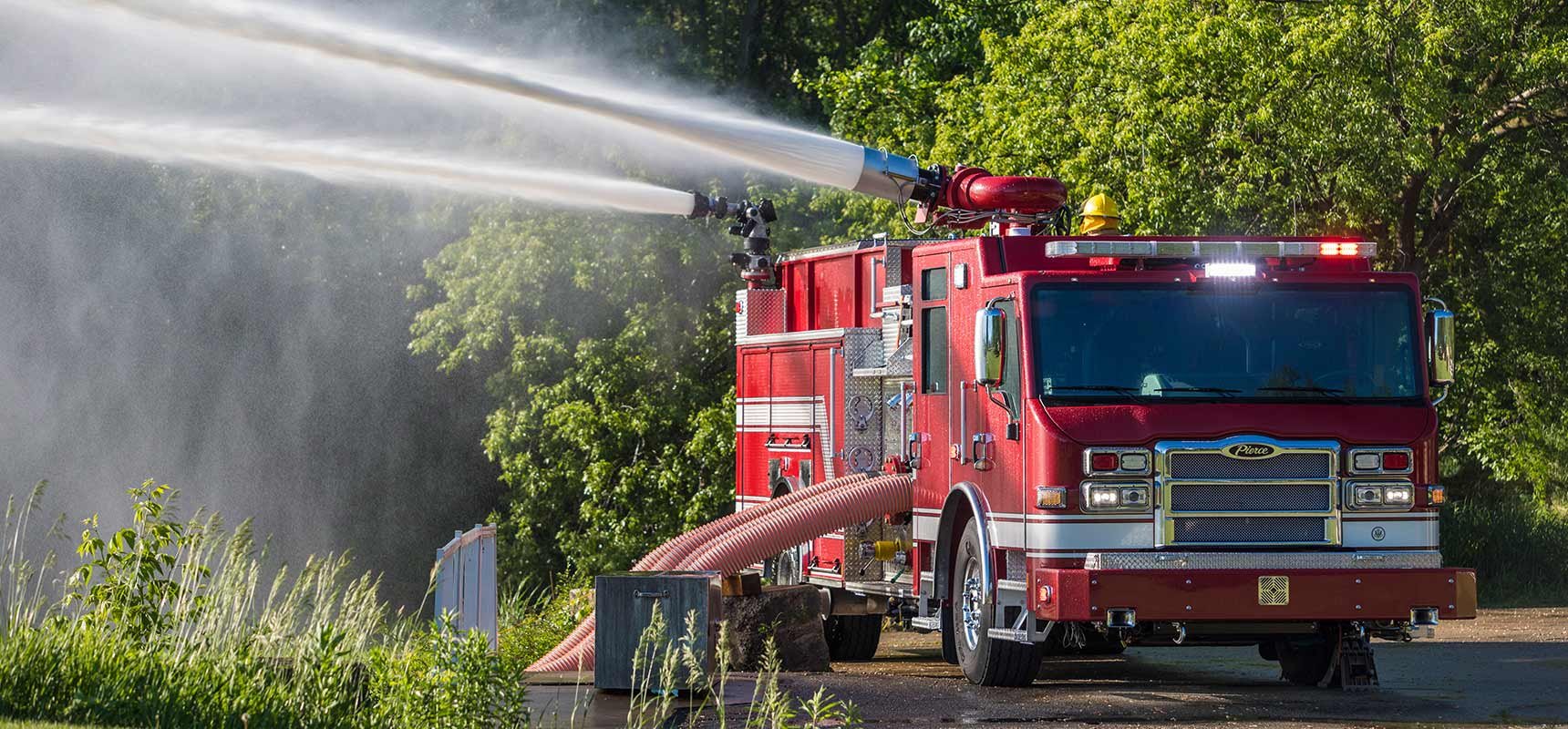 A red industrial fire truck is pumping water in a green wooded setting.