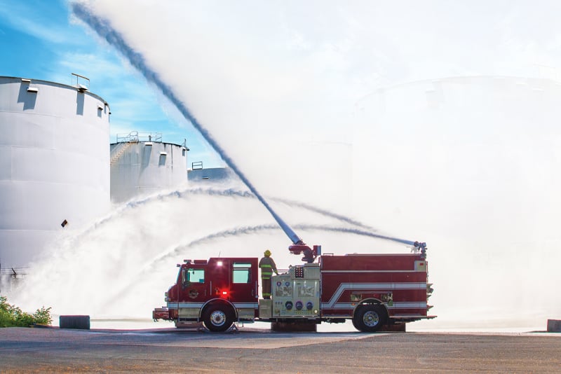 A red and while industrial fire truck is pumping water in the air at an oil refinery. 