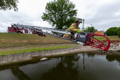 A ladder fire truck is lowered below-grade over water with a firefighter at the end practicing for a water rescue. 