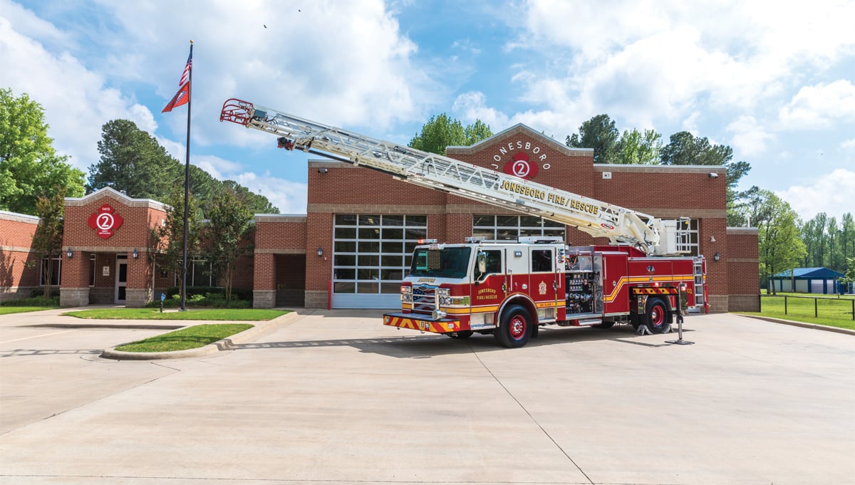 A red and white ladder fire truck is parked outside a fire station with the ladder raised and extended in the air. 