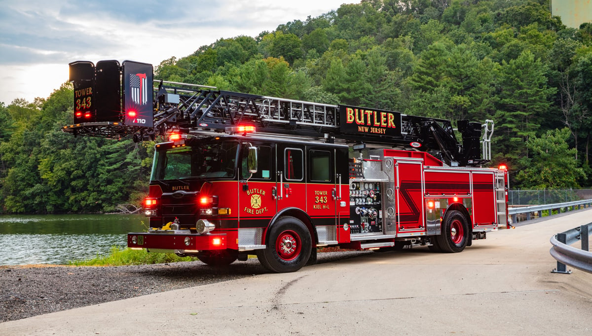 A red and black Pierce platform aerial fire truck is parked next to a wooded lake area. 