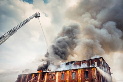 An aerial platform sprays water down as it towers above a large building that is on fire, spewing smoke and embers into the air. 