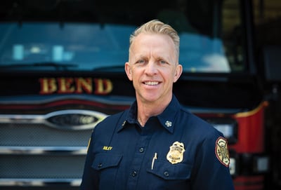 Bend Fire Chief, Todd Riley, in a blue uniform, standing in front of a fire apparatus.