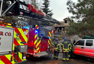Two fire trucks with lights on, one with the ladder extended, and a gray pickup truck, in front of a brown house.