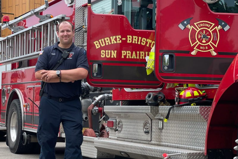 A Sun Prairie firefighter fire fighter stands outside a red fire truck outdoors.