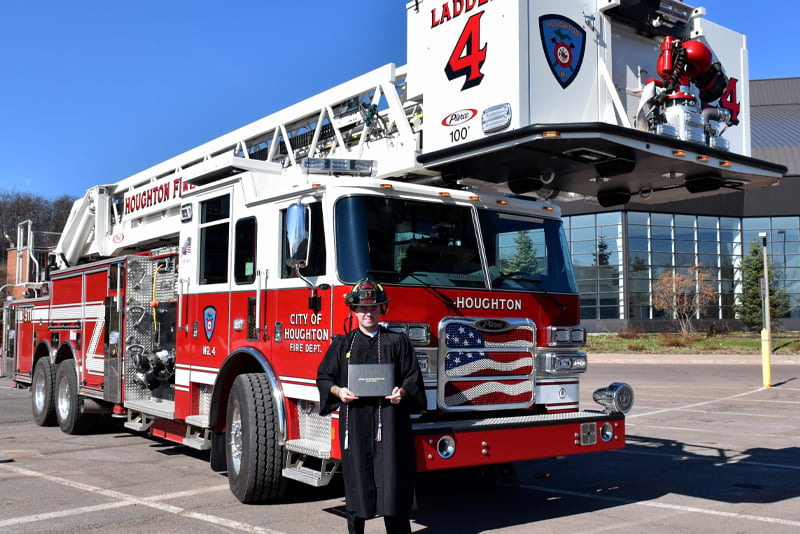 A volunteer firefighter stands in front of an aerial apparatus with a diploma.