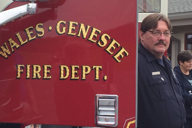 A volunteer firefighter stands next to the open door of a red fire truck.