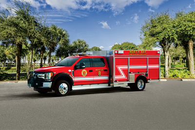 A red and white mini pumper fire apparatus is angle parked on an asphalt road with trees in the background.