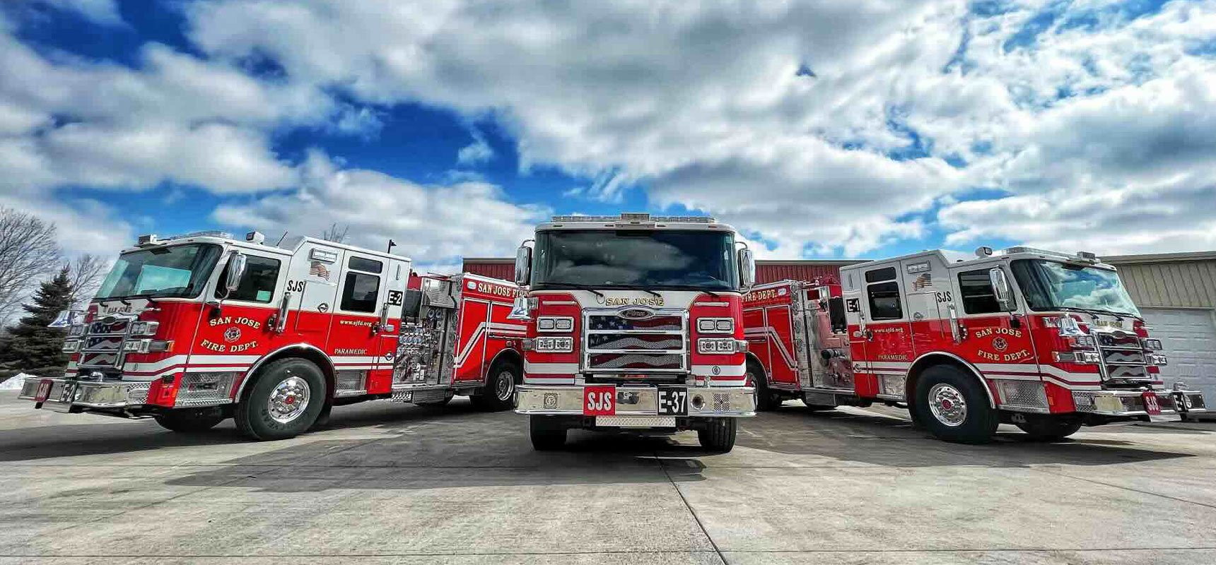Three pumper fire apparatus are parked outside of a warehouse facility featuring a bright blue sky scattered with white puffy clouds.