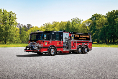 A red and black pumper tanker fire truck is parked on asphalt with green trees in the background. 