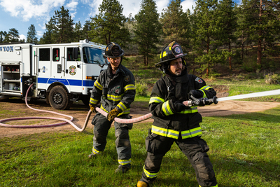 Two firefighters are holding a hose spraying water outside with a white firetruck and pine trees in the background.