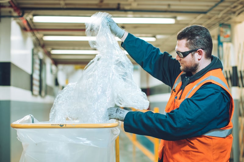 A man in an orange safety vests replaces a plastic bag in a recycling receptacle in a manufacturing facility. 