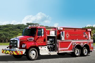 A red tanker fire truck is parked on asphalt with green rolling hills in the background and a blue sky scattered with puffy white clouds. 